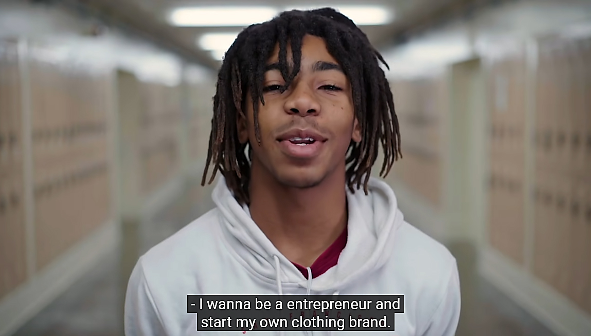 Portrait of Black, male, high school student in hallway with blurred lockers behind him.