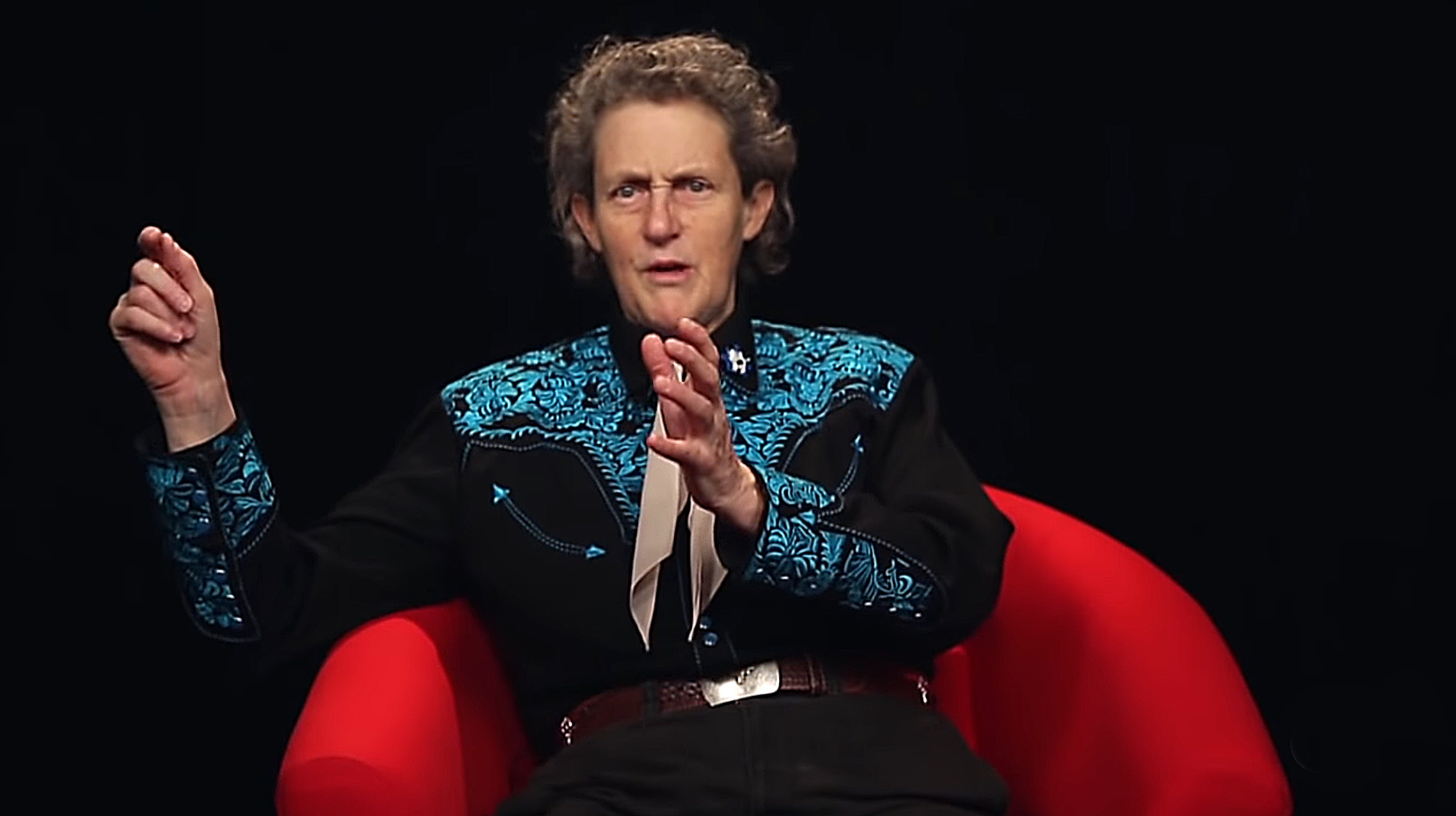Temple Grandin sitting on red couch wearing a black and blue western shirt and pants.