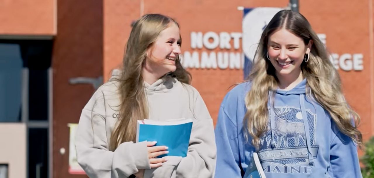 Two female students walk out of NSCC Lynn campus building chatting casually as they step into the sunlight