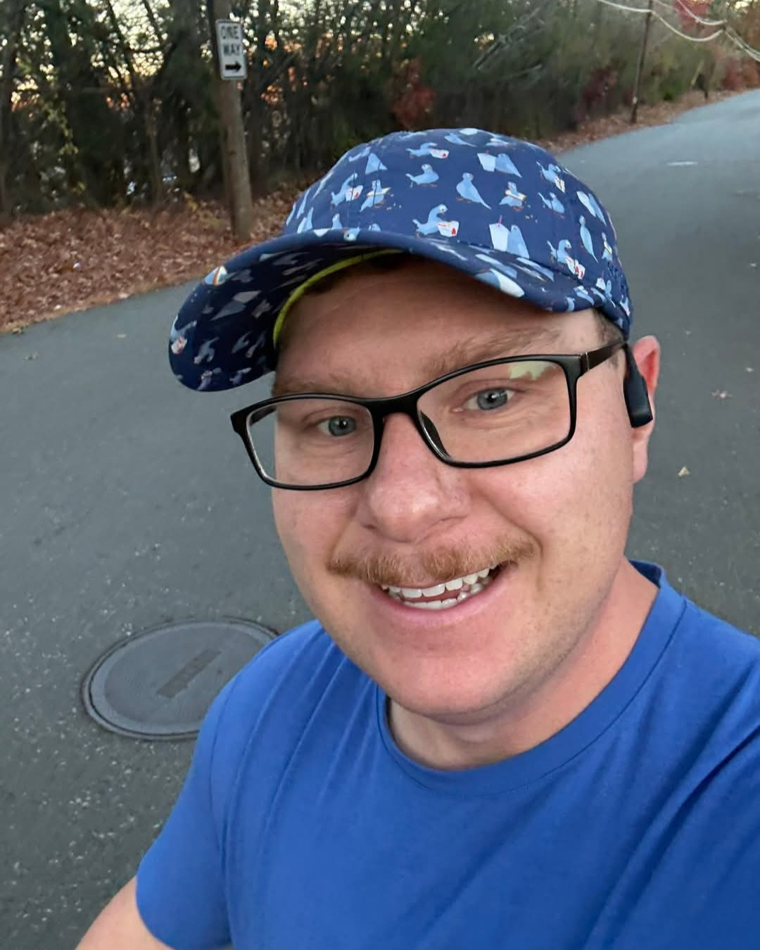 A man wearing glasses, a blue hat, and a shirt takes a selfie while running