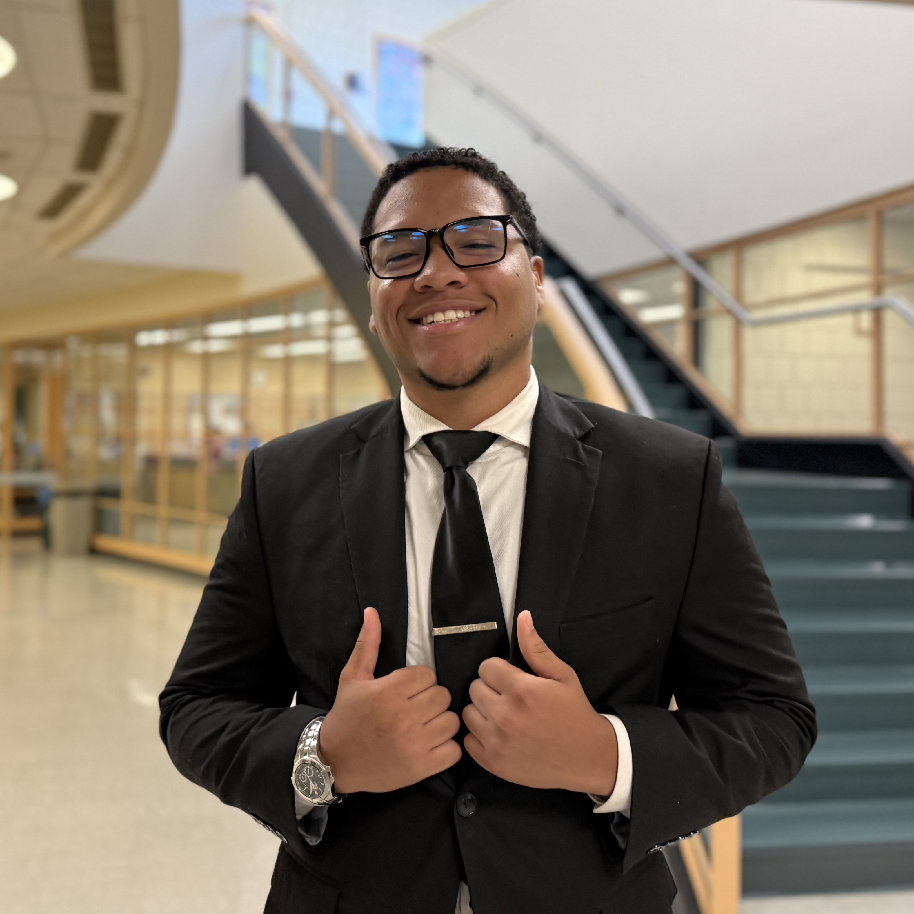 A person wearing glasses and a suit smiles at the camera with his hands on his lapels