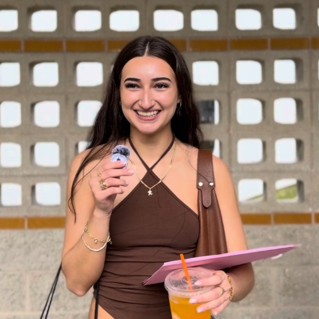 A person in a brown halterneck shirt smiles at the camera while holding a small microphone, a drink, and a folder