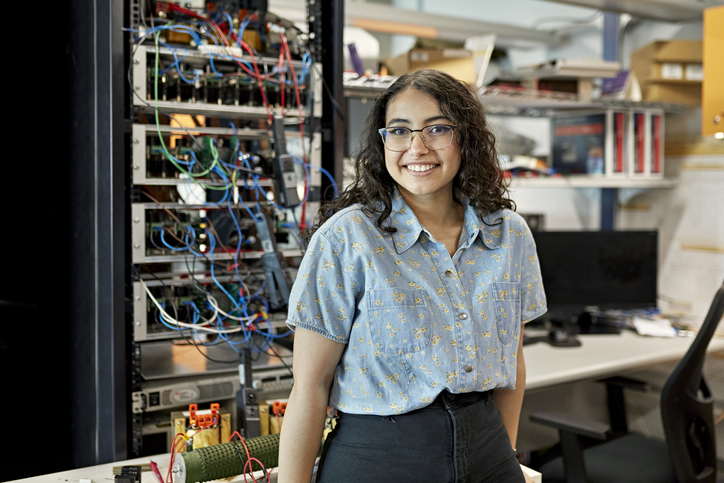 Waist-up view of casually dressed woman standing in electrical engineering lab and smiling at camera