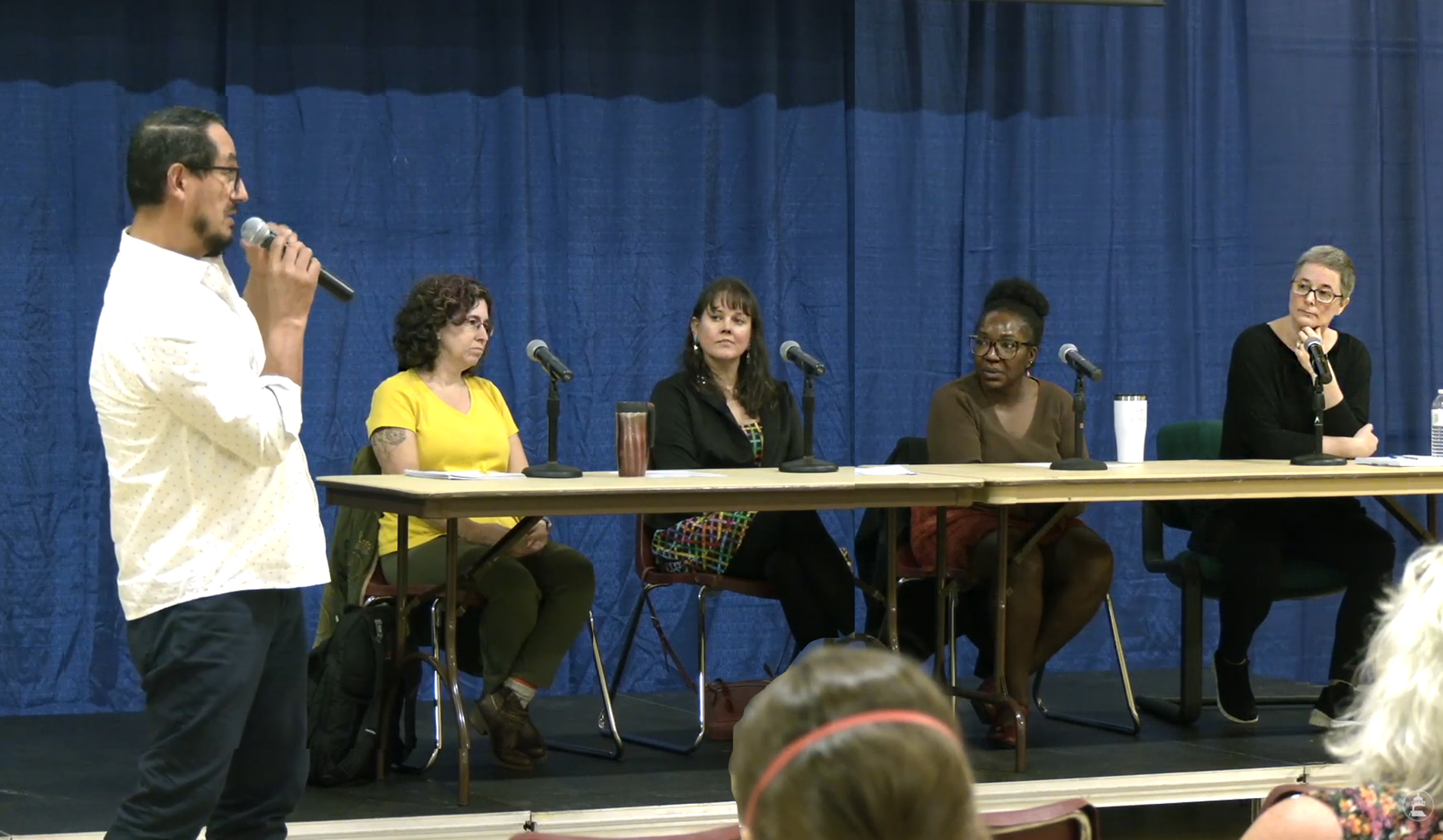 video still of four panelists at table with male questioner in foreground.